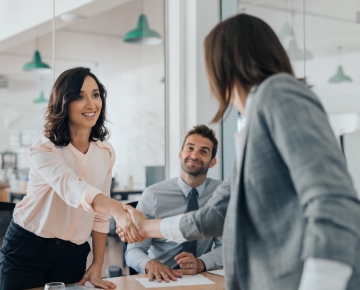 women shaking hands at desk next to man sitting in chair