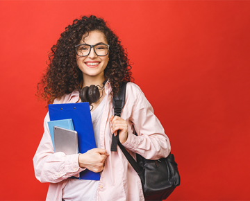 girl with curly hair and glasses wearing pink jacket with backpack and holding books against red background