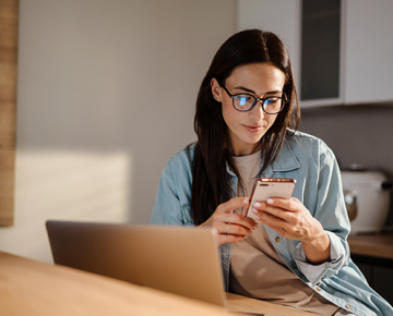 woman sitting at table looking at phone next to laptop